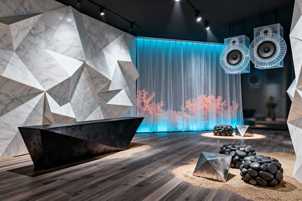 A media room features a dramatic faceted white marble wall beside a long angular blackened-steel bench. The dark wood floor holds circular patches of sand with pebble-like stone stools and small stainless-steel pyramids. At the back, sheer floor-to-ceiling curtains glow aqua with soft coral silhouettes, while two oversized transparent acrylic speakers hang from a black track-lit ceiling, casting focused light and reflections across the sculptural surfaces.
