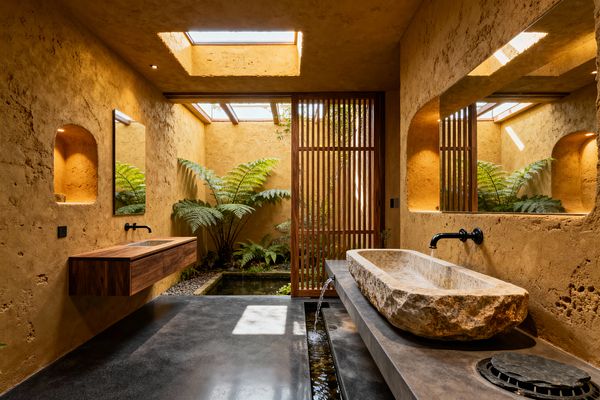 A skylit biophilic bathroom features textured ochre plaster walls, a rough stone trough sink set on a concrete counter, and matte black wall-mounted faucets. Lush ferns surround a shallow reflecting pond beside a cedar slatted screen, while a floating walnut vanity, arched wall niches with warm recessed lighting, river pebbles, wide mirrors, and a charcoal concrete floor create a serene, nature-connected space.
