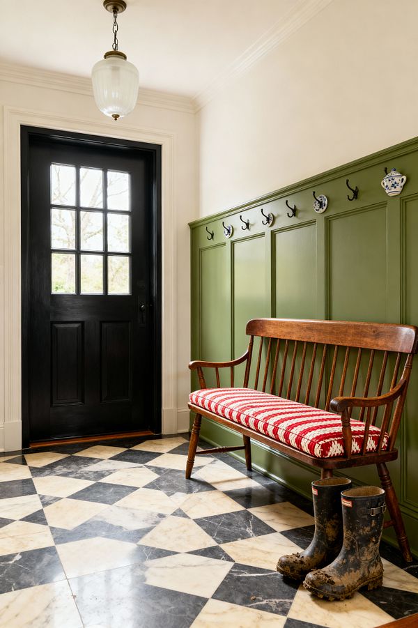 A traditional mudroom features a black, divided-light exterior door, cream walls, and olive green paneled wainscoting fitted with black metal hooks and blue-and-white ceramic pegs. A stained wood spindle-back bench with a red-and-white crocheted cushion lines the wall above a black-and-cream harlequin marble floor, while a milk-glass pendant hangs overhead and a pair of muddy rubber boots rest near the bench, signaling the space’s durable, welcoming purpose.