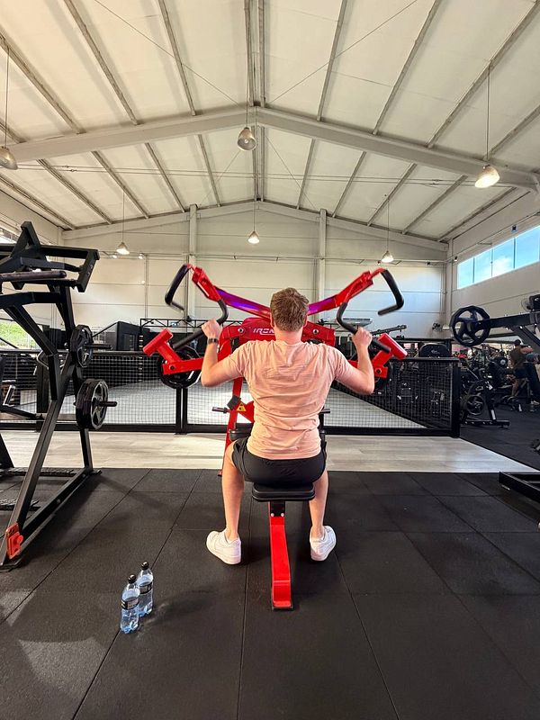 Interior view of a bright, modern gym with various workout equipment and a large window overlooking a city.