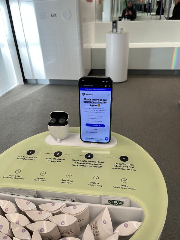 A smartphone displaying the Watchdog website is placed on a display stand featuring Pixel Buds in a Google store.