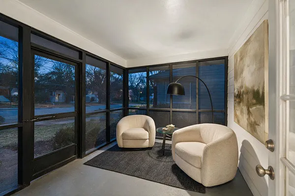 A modern screened front porch with black-framed glass walls, two cream rounded lounge chairs, a small glass side table, and a curved black floor lamp. A large abstract wall painting and a dark rug add texture against the white ceiling and light flooring.