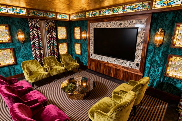 A maximalist media room with teal damask walls features two rows of velvet lounge chairs—chartreuse in front, magenta behind—facing a large flat-screen set in a terrazzo surround framed in walnut. Stained-glass wall panels and a clerestory band glow warmly alongside ribbed brass sconces. A circular smoked-glass coffee table sits on a taupe-and-charcoal striped carpet, with multicolored zigzag drapery and a reflective gold ceiling completing the layered look.