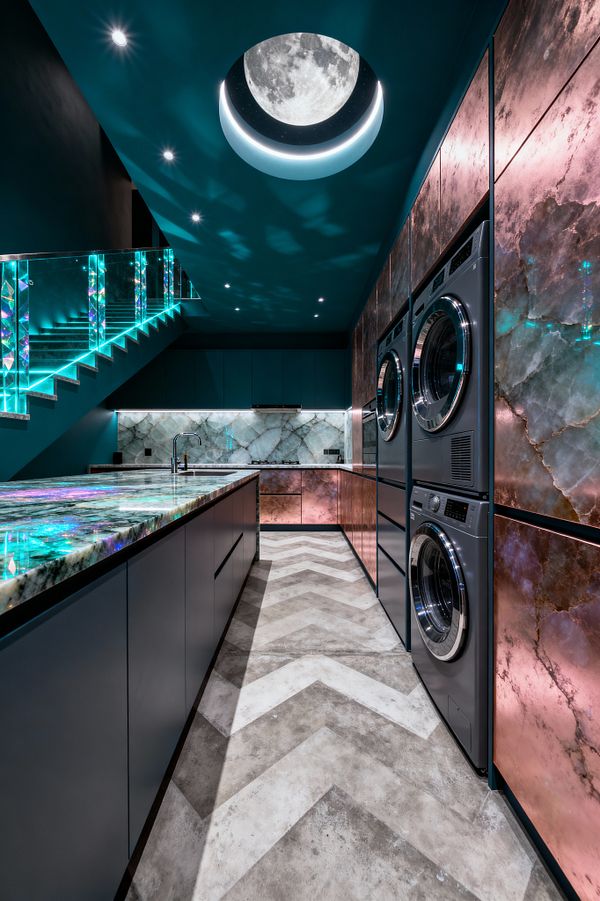 A narrow, futuristic laundry room features deep teal walls and a circular ceiling light that resembles a glowing full moon with a halo. Backlit iridescent stone countertops and rose-toned onyx cabinet panels frame stacked stainless-steel washers and dryers, while a glass stair balustrade edged in turquoise LEDs, a bookmatched gray marble backsplash, matte cabinetry, and a chevron concrete-look floor create a dramatic, cosmic ambience.
