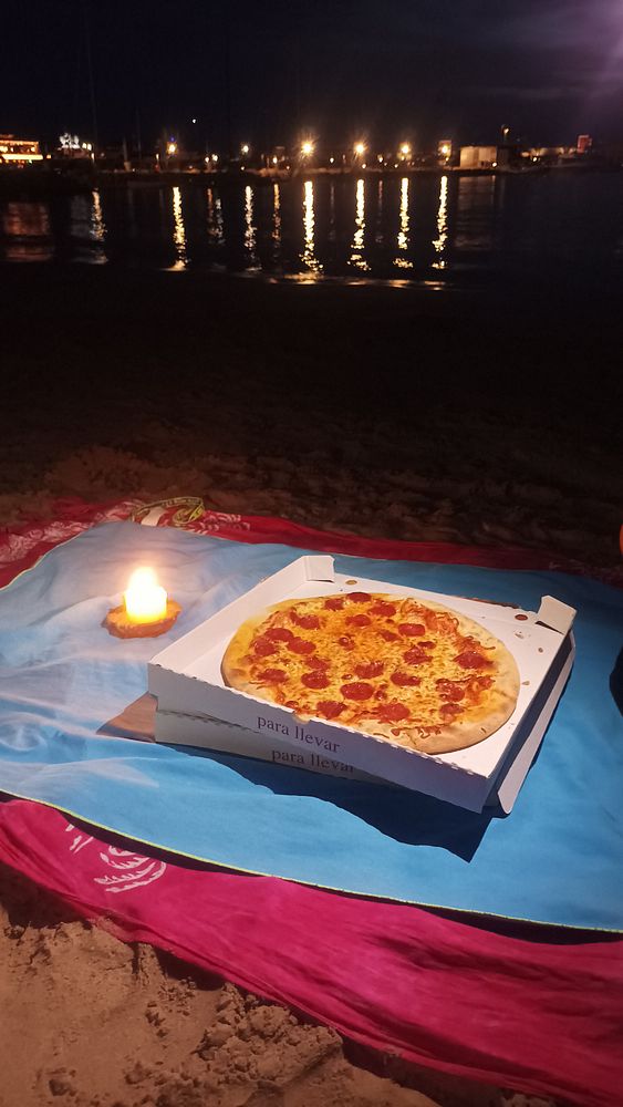 A pizza is placed on a beach blanket at night, illuminated by a candle and reflecting lights from the water.