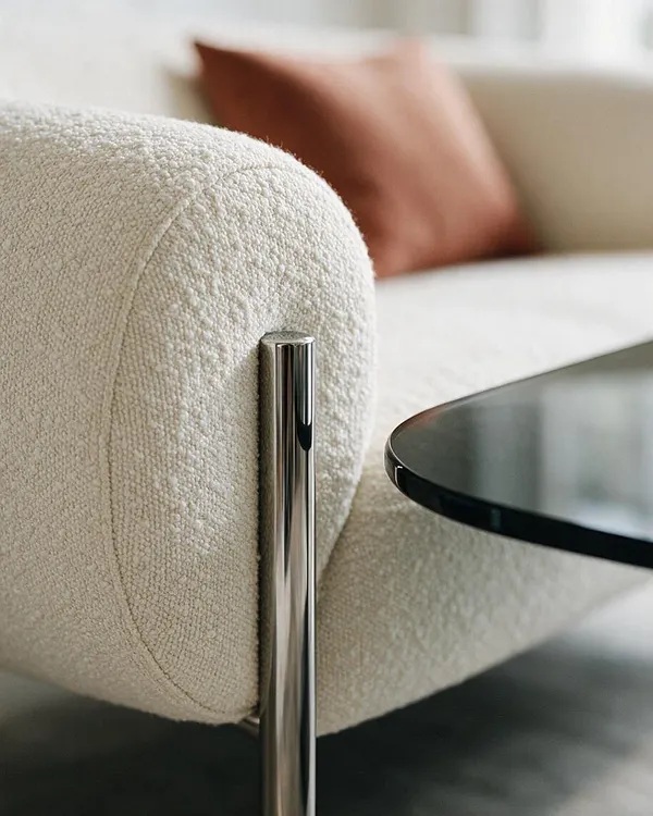 A close-up of a modern cream boucle sofa shows rounded upholstery and a polished chrome support leg beside a black glass side table. A soft brown cushion and light neutral seating create a calm, contemporary living room palette.