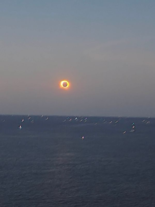 Photograph of a total solar eclipse showing the darkened sun with a bright, glowing corona against a dark sky above a silhouetted tree line.