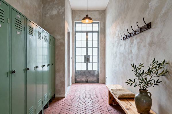 A narrow mudroom features terracotta herringbone brick flooring leading to a tall, gridded metal-and-glass French door illuminated by a single pendant. On the left is a row of sage-green metal lockers; on the right, a pale pine bench holds a woven cushion and a ceramic jug with olive branches beneath a line of dark iron hooks, all against softly mottled limewashed plaster walls for a rustic French Country atmosphere.