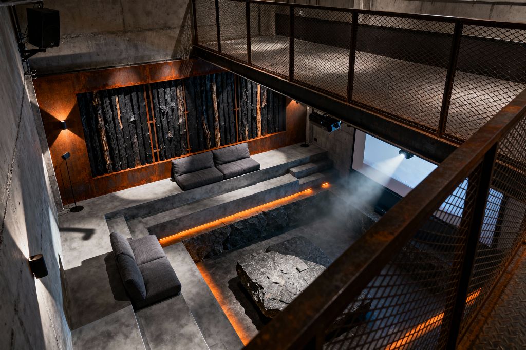 A double-height industrial media room with raw concrete walls, a sunken seating pit, and tiered concrete platforms with low gray sofas. A projector beams onto a wall screen; amber LED strips trace the pit’s edge around a rough-hewn stone table. A backdrop of Corten steel holds a charred-wood art panel, while a mesh metal mezzanine overlooks the space and black wall lights add focused illumination.