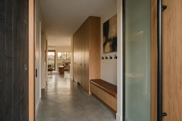 A modern entryway hallway with smooth concrete flooring, light oak built-in cabinetry, and a long wood bench with a brown cushioned seat. Black wall hooks and a framed abstract artwork add contrast, while a frosted glass door and a view into a bright living area complete the calm, streamlined space.