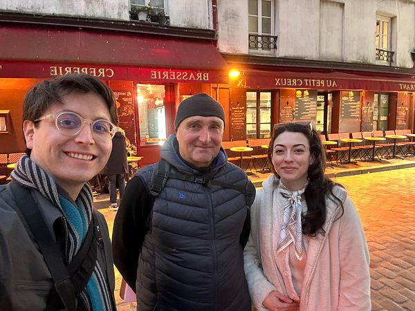 Three individuals pose together in a Parisian street with a café backdrop.