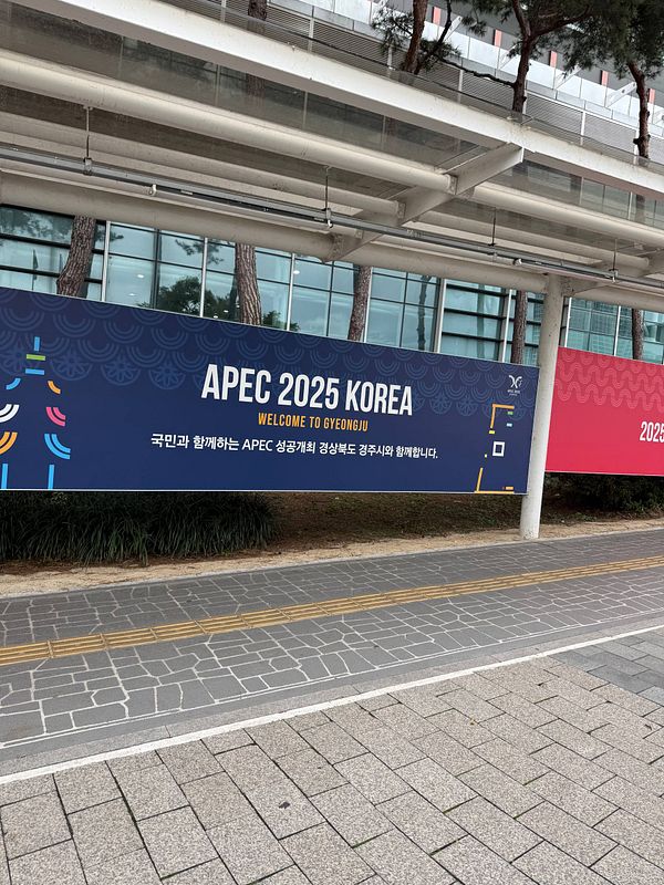 A large banner for APEC 2025 Korea welcoming visitors to Gyeongju is displayed outdoors by a building with glass windows and trees in the background.
