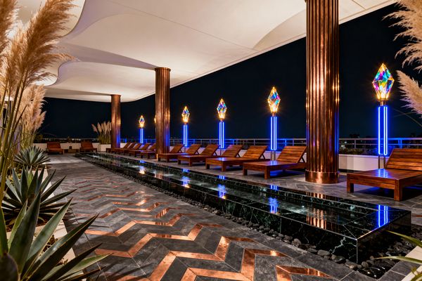 Nighttime rooftop patio featuring fluted copper columns supporting an undulating white canopy. A narrow black marble water feature with river rocks runs centrally, flanked by a chevron floor of dark stone with copper inlays. Slatted wooden loungers line one side, facing jewel-like torch lights on neon-blue glass columns. Agave and pampas grass planters soften the strong geometry and reflective surfaces in a richly lit Art Deco setting.