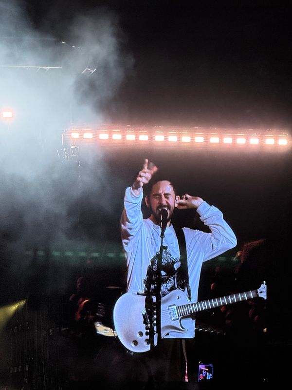 A musician performs on stage at a Linkin Park concert, surrounded by smoke and stage lights.