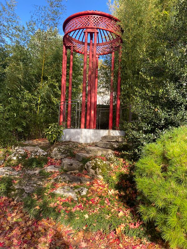 A red gazebo surrounded by lush greenery and fallen autumn leaves.