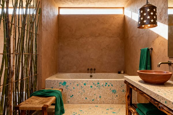 A Southwestern bathroom features adobe-toned plaster walls framing a built-in terrazzo soaking tub speckled with turquoise. At right, a terrazzo vanity holds a terracotta vessel sink with an aged bronze wall spout beneath a star-cut metal pendant; deep green towels, a woven rush bench, and a vertical screen of cane and cacti add texture, while a slim clerestory window washes the earthy finishes in warm desert light.