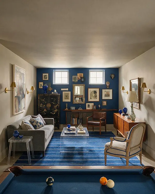 A long basement lounge with a blue accent wall at the far end, layered with framed artwork and small windows above. The room mixes a gray sofa, clear acrylic tables, a leather armchair, a wood sideboard, brass sconces, and a vivid blue striped rug, with a pool table in the foreground.