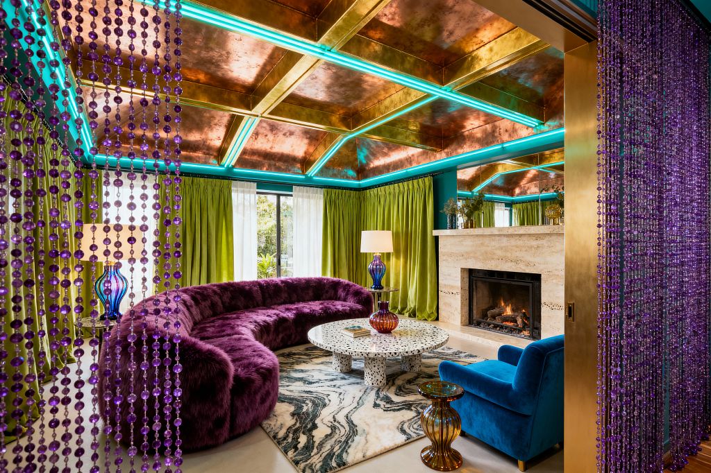 Vibrant maximalist living room with a metallic copper coffered ceiling accented by teal LED lighting, lime-green velvet curtains, and a travertine fireplace with a burning fire. A curved purple faux fur sectional wraps a round black-and-white terrazzo coffee table on a marbled rug. A cobalt blue velvet armchair and ribbed colored-glass lamps add shine. Purple beaded curtains frame the view, with large windows bringing in daylight.