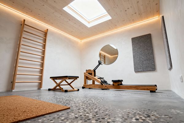 A minimal home gym features light wood stall bars, a wooden rowing machine, and a low folding bench with a black cushion, set against warm-white plaster walls. The space has a pine plank ceiling with a central skylight and warm LED cove lighting, a polished concrete floor with speckled pebble terrazzo, a round wall mirror, two gray felt acoustic panels, and a cork mat in the foreground.