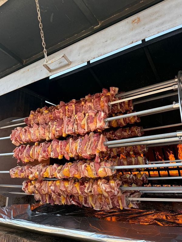 Photograph of a large outdoor barbecue grill loaded with multiple pieces of meat cooking over an open flame in a grassy area with a crowd and stage with lighting and speakers in the background.