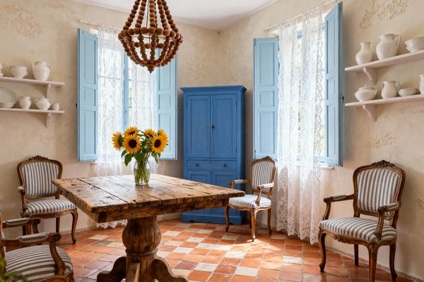 A French country dining room features a square reclaimed-wood pedestal table centered beneath a wooden bead chandelier. Terracotta checkerboard floor tiles and cream plaster walls set a warm backdrop for open shelves with white pottery, two tall windows dressed in lace curtains with sky-blue shutters, a matching blue painted armoire, and four carved Louis-style armchairs in blue-and-white ticking stripes; a clear glass vase of sunflowers sits on the table.
