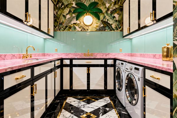 A glamorous U-shaped laundry room with black-and-cream cabinetry and brass pulls, topped by veined pink marble countertops. A mint-green back-painted glass backsplash runs the length of the walls, with undercabinet lighting. Tropical banana-leaf wallpaper and a round brass sconce with palm fronds crown the back wall. The floor is black-and-white marble with gold inlay, and a front-loading washer and dryer sit beneath the right counter.