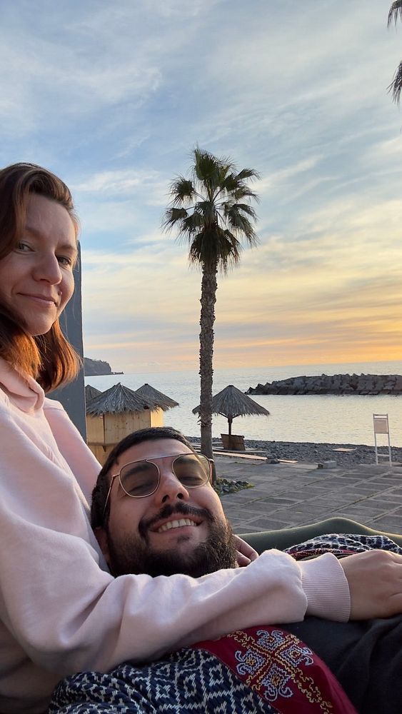 A couple enjoys a sunset by the beach, with palm trees and a serene ocean view in the background.