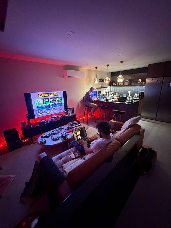 An evening coworking scene in a modern living room with two people working on laptops under warm pendant lights and a TV showing a retro game screen.