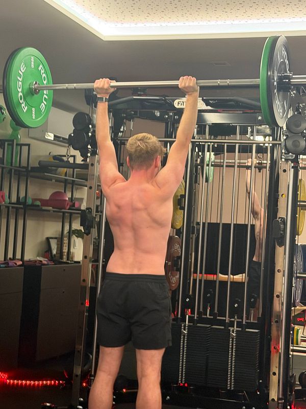 A shirtless man lifting a barbell with green Rogue weight plates overhead in a home gym.