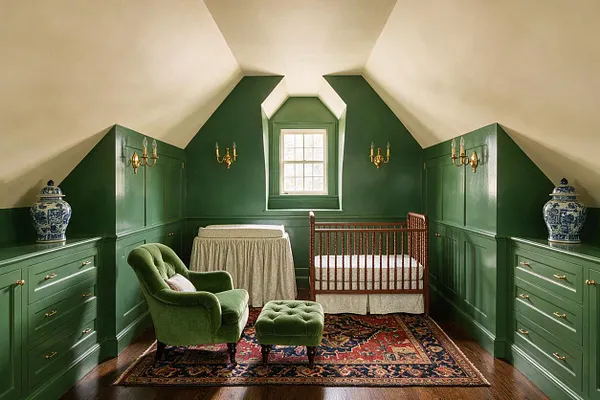 A traditional nursery in a sloped-ceiling room with deep green paneled walls and cream ceiling. The space includes a wooden crib, a draped changing table, a green velvet armchair and ottoman, brass wall sconces, blue-and-white ginger jars, and a patterned Persian-style rug over dark wood floors.