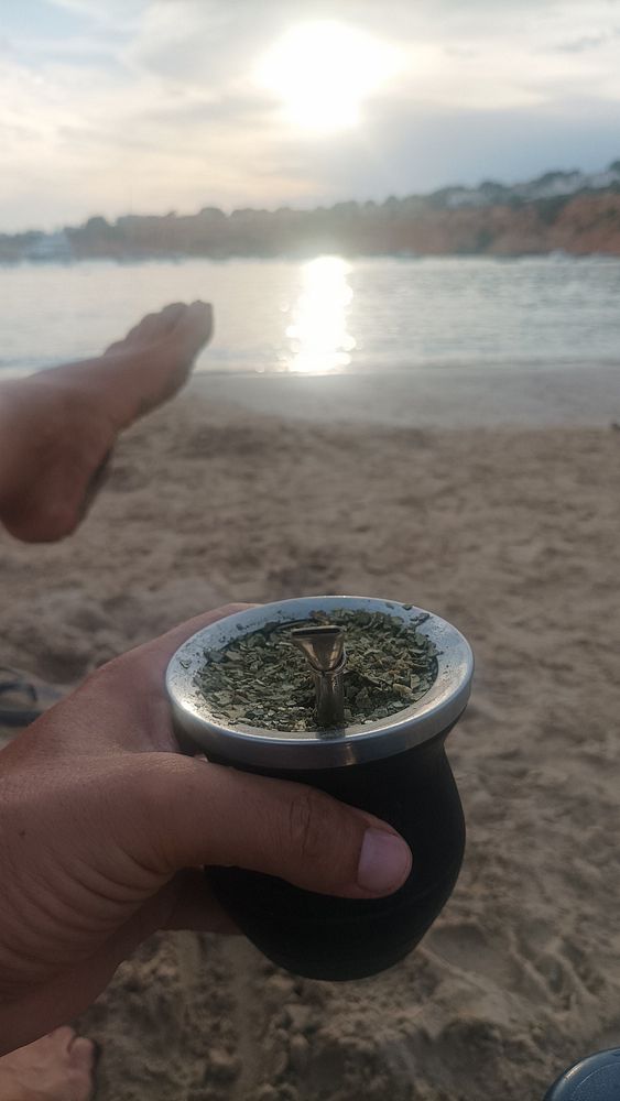 A hand holds a mate cup while a foot rests on the sandy beach, with a sunset reflecting on the water.