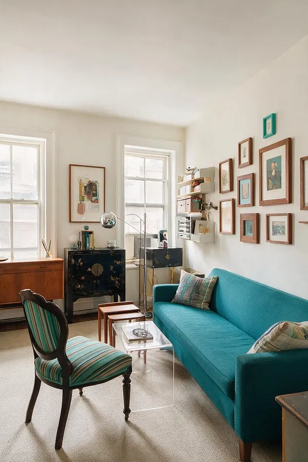A bright studio apartment living area with a turquoise sofa, a striped upholstered accent chair, and a clear acrylic coffee table. The walls are filled with framed artwork, and the room mixes vintage wood furniture, a black cabinet, and a brass floor lamp for an eclectic look.