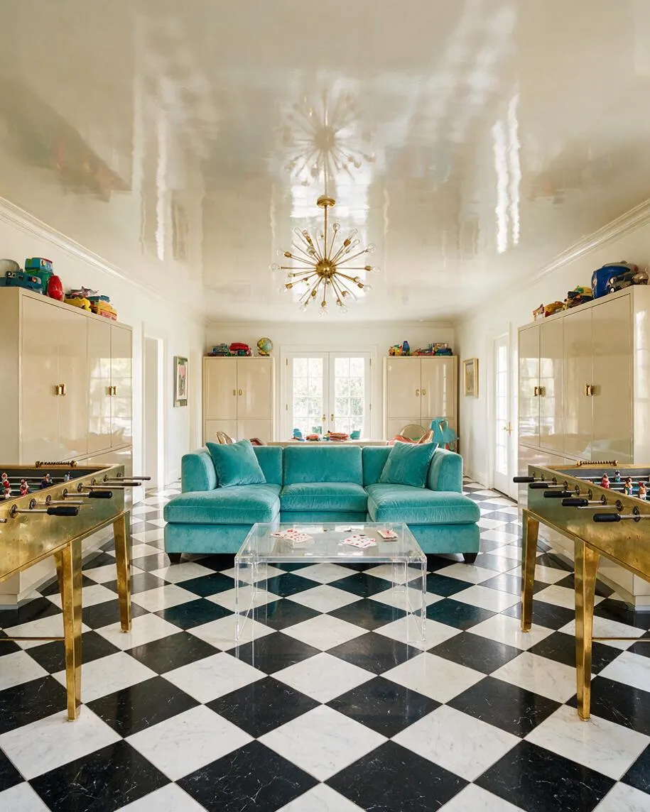 A bright game room in Hollywood Regency style features a black-and-white diamond marble floor, a glossy cream ceiling, and a teal velvet sectional centered in the room. Brass foosball tables, mirrored cabinetry, and a gold starburst chandelier add glamorous, playful contrast.