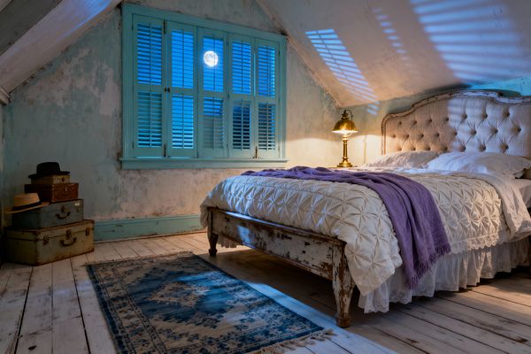 A cozy attic bedroom in shabby chic style shows a tufted linen headboard and a distressed wood bed dressed with a ruched white coverlet, lace bed skirt, and a lavender throw. Weathered white plank floors, peeling pale-blue walls, aqua plantation shutters framing a moonlit window, a faded blue rug, stacked vintage suitcases with hats, and a polished brass table lamp casting warm light create a romantic, timeworn atmosphere.