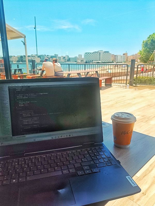 A laptop displaying code is set on a table next to a McCafé coffee cup, with a scenic view of a waterfront and people in the background.