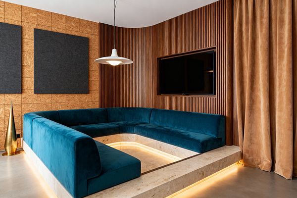 A mid-century modern media room features a sunken U-shaped teal velvet sofa set into a cream travertine platform with concealed warm LED lighting. A wall-mounted TV is framed by vertical walnut slats beside OSB wall tiles with large charcoal felt acoustic panels, while caramel velvet curtains, a smooth gray concrete floor, a white saucer pendant, and a small brass cone sculpture complete the space.