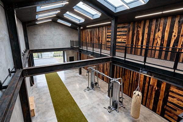 A double-height industrial home gym with polished concrete floors and exposed blackened steel beams. One wall is clad in vertical reclaimed wood planks with charred and honey tones, and a mezzanine catwalk with black mesh railing runs along it. Overhead skylights and linear LED fixtures brighten a green turf sprint lane, stainless-steel cable machines, and a hanging canvas punching bag beside a large opening to the exterior.