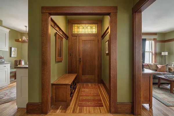 A Craftsman-style studio apartment entryway with thick natural wood trim, olive green walls, and a wooden door topped by a small stained-glass transom. A bench with cubbies sits along the hall, and the hardwood floor continues into adjacent living and kitchen areas.