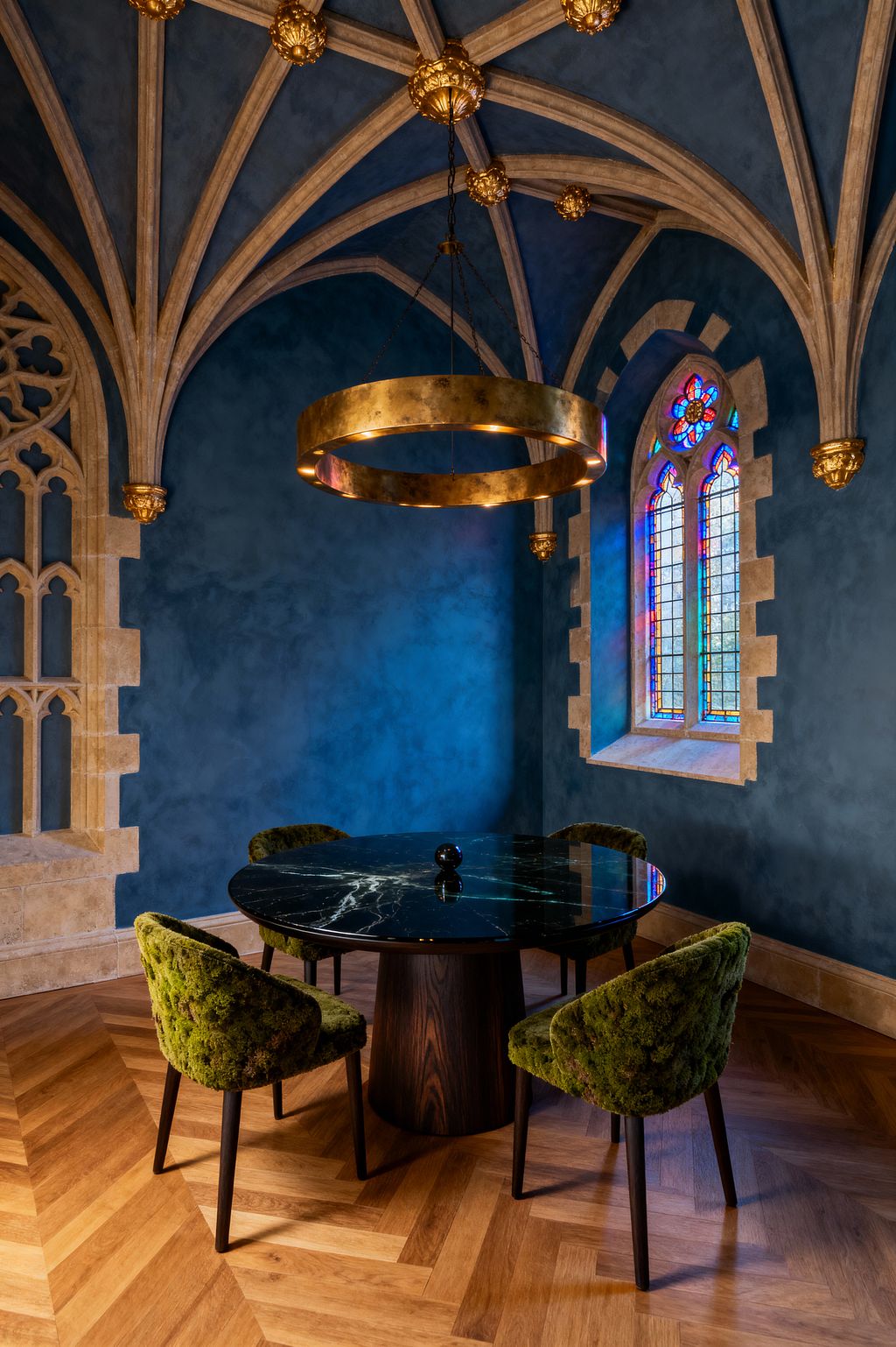 An intimate dining nook in a Gothic room with ribbed stone vaults, gilded bosses, and deep blue plastered walls. A large brass ring chandelier hangs over a round black marble table on a dark wood pedestal, surrounded by four moss‑green upholstered chairs. The floor is honey-toned oak in a herringbone pattern. A pointed lancet window with colorful stained glass and a limestone surround brings in filtered light.