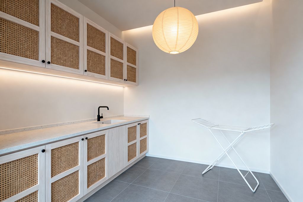 A minimalist Scandinavian laundry room with light wood cabinets featuring cane webbing panels runs along the left wall, topped by a pale quartz countertop and an integrated sink with a matte black faucet. Warm under-cabinet strip lighting and a ceiling cove glow illuminate white walls, while a round paper lantern hangs overhead; large gray floor tiles and a white folding drying rack keep the space calm and uncluttered.