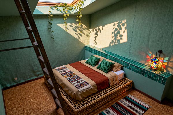 A compact Bohemian bedroom is lit by a skylight, featuring a low rattan platform bed layered with tan linen, a rust coverlet, a fringed kilim runner, and three green geometric pillows. A built-in ledge clad in glossy emerald tiles lines the wall, supporting a colorful stained-glass lantern that casts jewel-toned patterns; mint-textured walls, a trailing vine, cork flooring with a striped rug, and a rustic wooden ladder complete the sun-washed space.