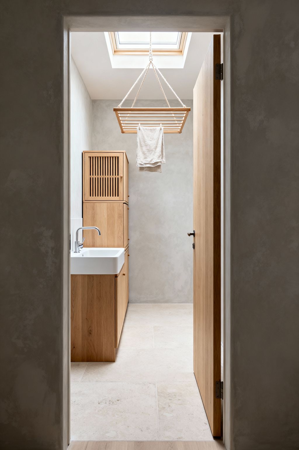 A narrow Scandinavian-style laundry room with light troweled plaster walls and large pale stone tiles, seen through an open oak door. Oak cabinets line the left side with a white rectangular ceramic sink and chrome tap, while a slatted wooden ceiling airer hangs from thick ropes beneath a skylight, drying a single linen towel. The soft neutral palette highlights natural materials, clean lines, and ample daylight.