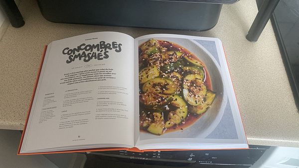 A recipe book opened to a page featuring a dish called 'Concombres Smashés' with a vibrant cucumber salad.