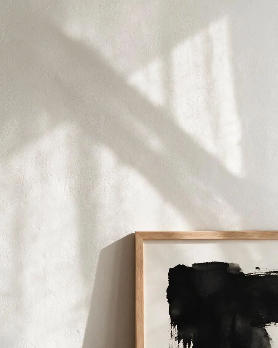 A textured warm white wall lit by soft natural shadows, with the top left of a framed abstract ink artwork visible in the lower right. The artwork has a light natural oak frame and a bold black brushstroke on white paper, creating a calm Japandi-inspired composition.