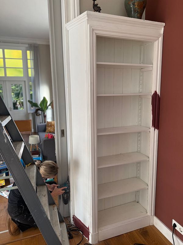 A person working on painting and sanding a white bookshelf and adjacent wall corner in a room with wooden flooring.