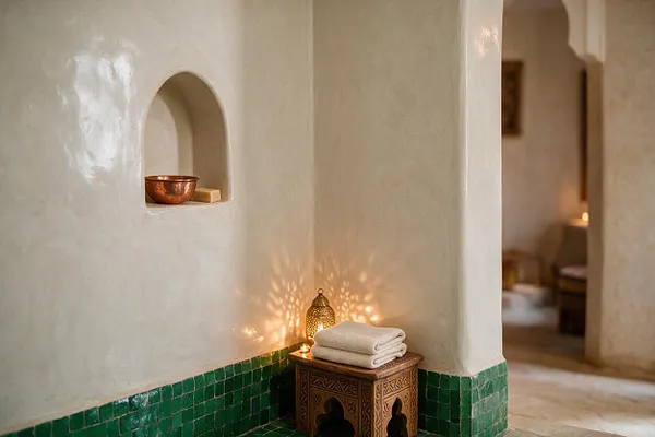 A serene Moroccan-style interior with smooth cream plaster walls, a recessed arched niche holding a copper bowl, and a low carved wooden stool topped with folded white towels. Green zellige tile wraps the base of the walls, while a brass lantern casts patterned light across the corner and warm arches lead into a softly lit adjacent room.