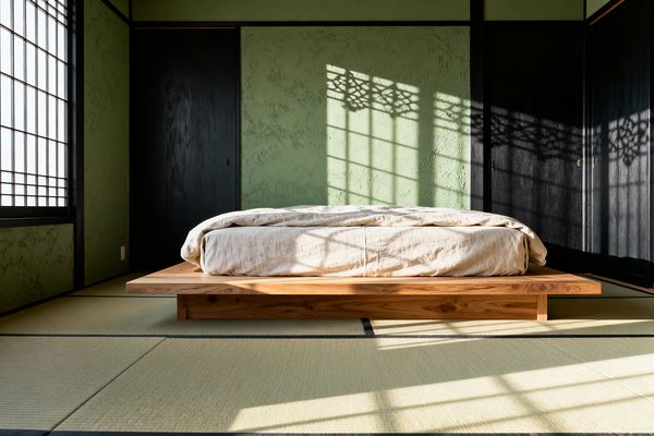 A minimalist Japanese bedroom centers a low solid wood platform bed with off-white linen on woven tatami mats. Textured sage-green plaster walls are articulated by black timber framing and sliding doors. At left, a shoji window filters sunlight, projecting geometric lattice shadows across the wall and floor. The room is uncluttered and symmetrical, emphasizing natural materials, muted colors, and calm proportions that evoke a Zen retreat.