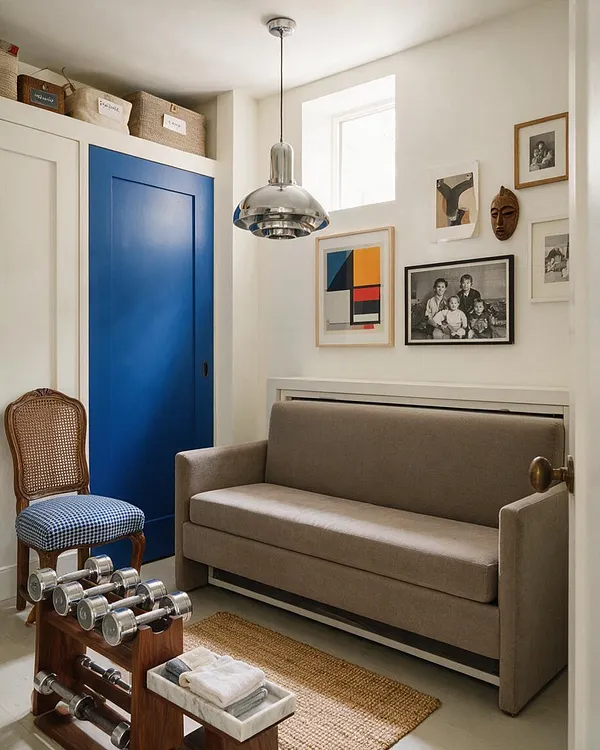 A compact home gym doubles as a stylish sitting area, with a taupe sofa, a blue sliding door, a cane-back chair with a patterned seat, and a wooden dumbbell rack in the foreground. Framed artwork, a chrome pendant light, woven storage baskets, and a natural jute rug add eclectic character against white walls.