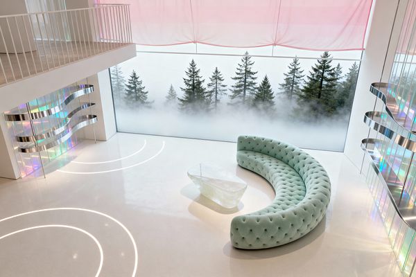 A double-height living room with a vast floor-to-ceiling window overlooking fog-shrouded evergreens features a curved, tufted mint-green sofa around a translucent sculptural coffee table. The space has a seamless white floor with concentric LED arcs, iridescent wall panels with wave-like stainless-steel bands, a white mezzanine with slim railings, and a pale pink sheer shade diffusing daylight.