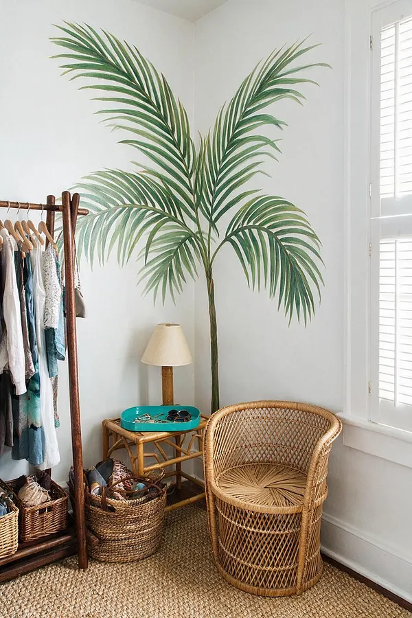 A bright closet corner features a large painted palm tree mural on white walls, a woven rattan chair, baskets, and a bamboo-style side table with a small lamp. A wooden clothing rack with hanging garments sits beside natural fiber flooring and a window with white shutters.